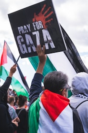 Several people are gathered at what appears to be a protest or rally. One person in the foreground is draped in a Palestinian flag and holding a sign that reads 'PRAY FOR GAZA' with a handprint on it. Other demonstrators and flags can be seen in the background.