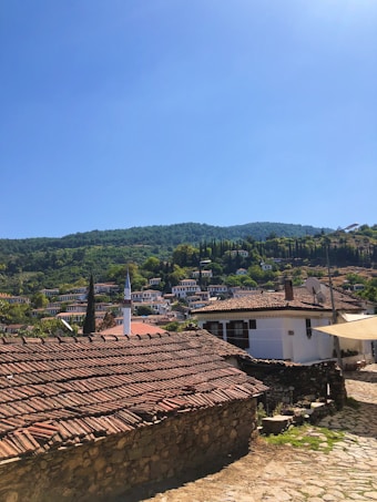 A quaint village with traditional houses featuring red-tiled roofs nestled into a hillside. A white minaret rises among the homes, surrounded by lush greenery and topped with a vibrant blue sky.