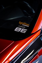 A close-up view of the corner of a car with a sleek, red finish and carbon fiber detailing. A decal is visible on the window with the text 'Ignition Auto Repairing' in orange and '86' in white. The car's glossy surface reflects light, adding to its dynamic and sporty aesthetic.
