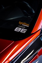 A close-up view of the corner of a car with a sleek, red finish and carbon fiber detailing. A decal is visible on the window with the text 'Ignition Auto Repairing' in orange and '86' in white. The car's glossy surface reflects light, adding to its dynamic and sporty aesthetic.
