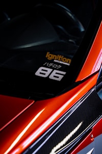 A close-up view of the corner of a car with a sleek, red finish and carbon fiber detailing. A decal is visible on the window with the text 'Ignition Auto Repairing' in orange and '86' in white. The car's glossy surface reflects light, adding to its dynamic and sporty aesthetic.