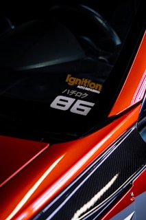 A close-up view of the corner of a car with a sleek, red finish and carbon fiber detailing. A decal is visible on the window with the text 'Ignition Auto Repairing' in orange and '86' in white. The car's glossy surface reflects light, adding to its dynamic and sporty aesthetic.