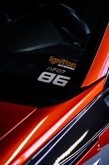 A close-up view of the corner of a car with a sleek, red finish and carbon fiber detailing. A decal is visible on the window with the text 'Ignition Auto Repairing' in orange and '86' in white. The car's glossy surface reflects light, adding to its dynamic and sporty aesthetic.