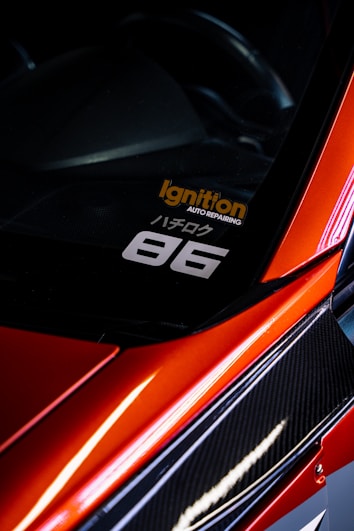 A close-up view of the corner of a car with a sleek, red finish and carbon fiber detailing. A decal is visible on the window with the text 'Ignition Auto Repairing' in orange and '86' in white. The car's glossy surface reflects light, adding to its dynamic and sporty aesthetic.