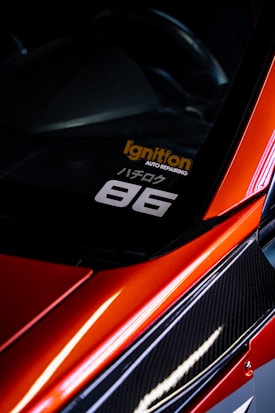 A close-up view of the corner of a car with a sleek, red finish and carbon fiber detailing. A decal is visible on the window with the text 'Ignition Auto Repairing' in orange and '86' in white. The car's glossy surface reflects light, adding to its dynamic and sporty aesthetic.
