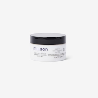 A matte texturizing paste container is centered in the image. The cylindrical container features a white body with a black lid. The brand name 'Milbon' is prominently displayed, along with product details in blue and grey text.