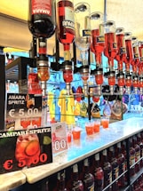 A row of liquor bottles with spigots, dispensing bright red drinks into plastic cups on a countertop. Several signs advertise Amara Spritz and Campari Spritz for €5.00. In the background, colorful clothing, including shirts and jackets, are visible hanging on racks.