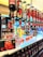 A row of liquor bottles with spigots, dispensing bright red drinks into plastic cups on a countertop. Several signs advertise Amara Spritz and Campari Spritz for €5.00. In the background, colorful clothing, including shirts and jackets, are visible hanging on racks.