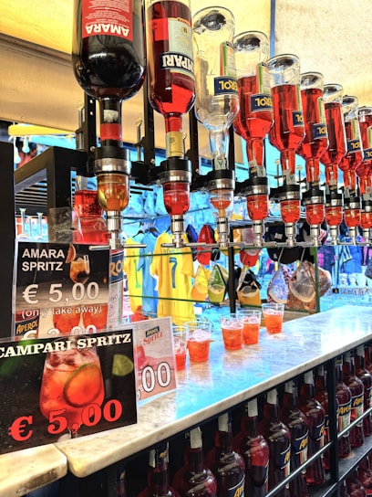 A row of liquor bottles with spigots, dispensing bright red drinks into plastic cups on a countertop. Several signs advertise Amara Spritz and Campari Spritz for €5.00. In the background, colorful clothing, including shirts and jackets, are visible hanging on racks.