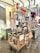 A vibrant street food cart displays a variety of drinks, including Aperol Spritz, with several upside-down alcohol bottles. The cart is decorated with colorful, traditional Italian art. Nearby, a stall sells souvenirs, including magnets and clothes with a Western theme. The setting is under a tent or canopy, which provides shade in an open-air market area.