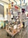 A vibrant street food cart displays a variety of drinks, including Aperol Spritz, with several upside-down alcohol bottles. The cart is decorated with colorful, traditional Italian art. Nearby, a stall sells souvenirs, including magnets and clothes with a Western theme. The setting is under a tent or canopy, which provides shade in an open-air market area.