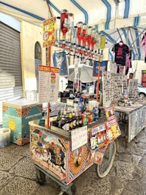 A vibrant street food cart displays a variety of drinks, including Aperol Spritz, with several upside-down alcohol bottles. The cart is decorated with colorful, traditional Italian art. Nearby, a stall sells souvenirs, including magnets and clothes with a Western theme. The setting is under a tent or canopy, which provides shade in an open-air market area.