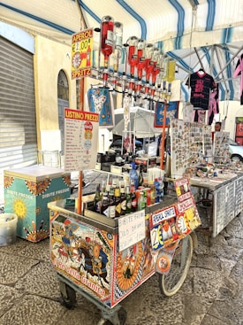 A vibrant street food cart displays a variety of drinks, including Aperol Spritz, with several upside-down alcohol bottles. The cart is decorated with colorful, traditional Italian art. Nearby, a stall sells souvenirs, including magnets and clothes with a Western theme. The setting is under a tent or canopy, which provides shade in an open-air market area.