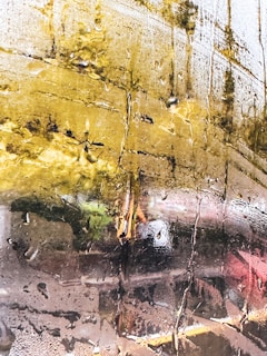 A close-up view of a glass surface covered in condensation, with streaks of water droplets creating a textured pattern. Reflections of a blurred outdoor scene, possibly a landscape with trees and greenery, are visible through the glass, giving a distorted view.