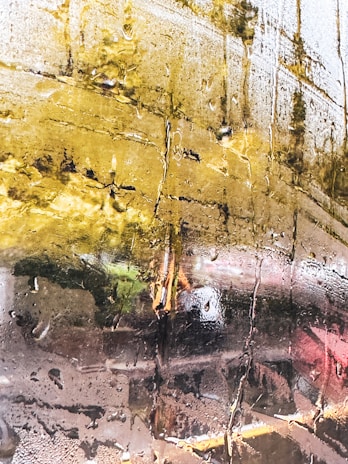 A close-up view of a glass surface covered in condensation, with streaks of water droplets creating a textured pattern. Reflections of a blurred outdoor scene, possibly a landscape with trees and greenery, are visible through the glass, giving a distorted view.