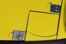 The image features a yellow panel with two metal fasteners. Each fastener is connected by a wire loop, appearing to secure the panel in place. The surface is glossy, and there is a line down the middle suggesting a compartment or cover.