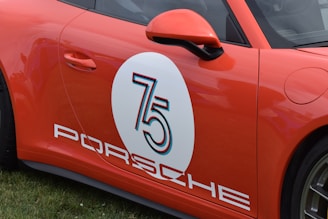 A bright orange car with a large circular white decal on the side displaying the number 75, featuring stylized text and the word 'PORSCHE' below it.