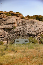 A white caravan is situated in a natural setting, surrounded by tall grass and positioned in front of large rocky formations. Sparse trees and bushes are scattered around the area, with a blue sky overhead. The landscape appears rugged and untouched, giving a sense of isolation.