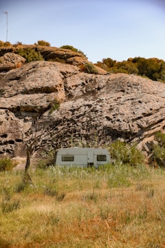 A white caravan is situated in a natural setting, surrounded by tall grass and positioned in front of large rocky formations. Sparse trees and bushes are scattered around the area, with a blue sky overhead. The landscape appears rugged and untouched, giving a sense of isolation.
