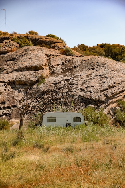A white caravan is situated in a natural setting, surrounded by tall grass and positioned in front of large rocky formations. Sparse trees and bushes are scattered around the area, with a blue sky overhead. The landscape appears rugged and untouched, giving a sense of isolation.