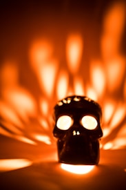 A black skull-shaped object appears prominently in the center, with bright lights emanating from its eyes and casting intricate patterns of light and shadow around it. The warm hues of orange and yellow dominate the background, creating an intriguing atmospheric effect.