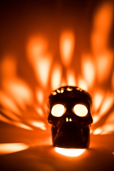 A black skull-shaped object appears prominently in the center, with bright lights emanating from its eyes and casting intricate patterns of light and shadow around it. The warm hues of orange and yellow dominate the background, creating an intriguing atmospheric effect.