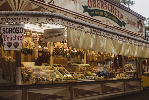 A festive market stall offering a variety of sweets and candies, including intricately decorated gingerbread hearts hanging from the top. The signage announces products like 'Zuckerwatte' and 'Schoko Früchte' suggesting cotton candy and chocolate fruits. The stall is adorned with colorful confections neatly displayed in containers and jars. The atmosphere appears warm and inviting.