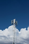 A tall communication tower stands against a backdrop of fluffy white clouds and a clear blue sky.