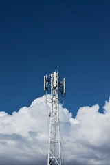 A tall communication tower stands against a backdrop of fluffy white clouds and a clear blue sky.