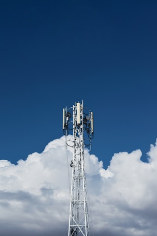 A tall communication tower stands against a backdrop of fluffy white clouds and a clear blue sky.