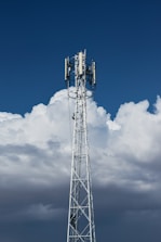 A tall, metal telecommunications tower stands against a backdrop of dense, white clouds and a deep blue sky. The structure is equipped with various electronic components, including antennae and cables.