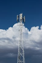 A tall, metal telecommunications tower stands against a backdrop of dense, white clouds and a deep blue sky. The structure is equipped with various electronic components, including antennae and cables.