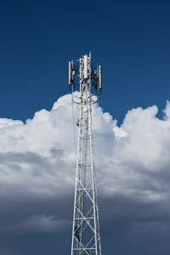 A tall, metal telecommunications tower stands against a backdrop of dense, white clouds and a deep blue sky. The structure is equipped with various electronic components, including antennae and cables.