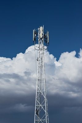 A tall, metal telecommunications tower stands against a backdrop of dense, white clouds and a deep blue sky. The structure is equipped with various electronic components, including antennae and cables.