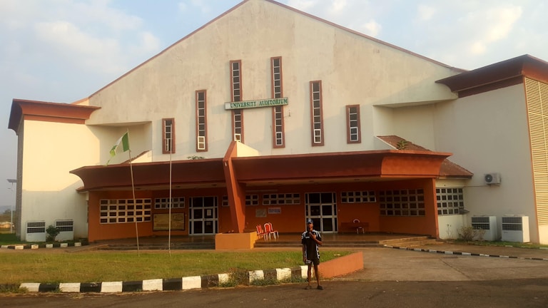 A large building with a sign that reads 'University Auditorium' is shown. The architecture includes a white and red-brown color scheme, with several narrow windows running vertically along the front. There is a small, grassy area in front of the building, with a walkway leading to the entrance. A flagpole with a green and white flag is visible, and a person stands in front of the building.