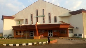 A large building with a sign that reads 'University Auditorium' is shown. The architecture includes a white and red-brown color scheme, with several narrow windows running vertically along the front. There is a small, grassy area in front of the building, with a walkway leading to the entrance. A flagpole with a green and white flag is visible, and a person stands in front of the building.