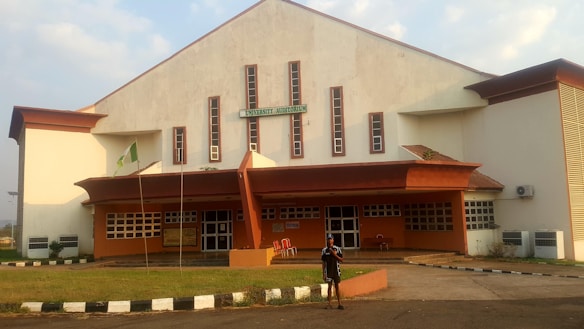 A large building with a sign that reads 'University Auditorium' is shown. The architecture includes a white and red-brown color scheme, with several narrow windows running vertically along the front. There is a small, grassy area in front of the building, with a walkway leading to the entrance. A flagpole with a green and white flag is visible, and a person stands in front of the building.