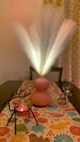 A decorative table setup featuring a pink vase with illuminated white pampas grass as the centerpiece. Two candle holders shaped like reindeer with red candles are placed on each side. The table is draped with a colorful, patterned runner displaying various floral and abstract designs. In the background, patterned curtains complement the aesthetic.