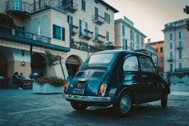 A vintage car is parked in a quaint European square surrounded by traditional buildings. The architecture features balconies with plants and arched doorways. A few people are sitting at an outdoor caf&eacute;, enjoying their time, creating a serene, leisurely atmosphere.