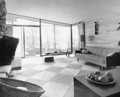 A mid-century modern living room featuring a sectional sofa with cushions, a wooden coffee table, and a decorative bowl on a side table. The room has large windows with sheer curtains allowing natural light to flood in, and a textured wall with a decorative piece. There are elements of natural materials like wood and stone.