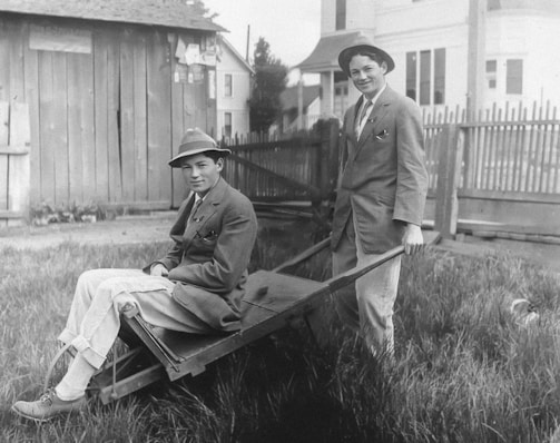Two young men in formal suits and hats are outdoors. One sits on a wooden wheelbarrow while the other stands behind it, holding the handles, with a playful expression. They are in a grassy yard with a wooden fence and buildings in the background, suggesting a residential area.