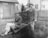Two young men in formal suits and hats are outdoors. One sits on a wooden wheelbarrow while the other stands behind it, holding the handles, with a playful expression. They are in a grassy yard with a wooden fence and buildings in the background, suggesting a residential area.