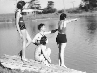 Four people wearing vintage swimsuits are on a log by the water, with two of them pointing towards something in the distance. The setting is calm, with a backdrop of trees and a serene body of water.