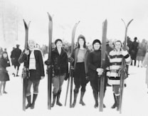 A group of five women dressed in early 20th-century ski attire stand side by side holding skis. They are wearing winter clothing including boots, coats, and hats. Behind them, a gathering of people in winter clothes can be seen in a snowy outdoor setting.