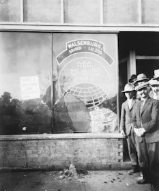 A historical black and white photograph featuring a group of men in suits and hats standing beside a window of a building. The window displays signage for the Walsenburg Branch I.U. 220 of the Industrial Workers of the World (I.W.W.), with visible damage such as cracks and a hole. A sign in the window says 'Now is vacation time, live larger.' The figures in the photo look towards the camera with serious expressions. Below the window, a brick wall is visible, with rubble on the ground.