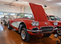 A vintage red convertible car with its hood open is prominently displayed in a well-lit showroom. The car features chrome detailing and classic design elements characteristic of mid-20th century vehicles. It is surrounded by other cars in the background, also appearing to be classic models. The showroom has wooden flooring and large windows that let in natural light.