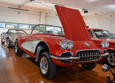 A vintage red convertible car with its hood open is prominently displayed in a well-lit showroom. The car features chrome detailing and classic design elements characteristic of mid-20th century vehicles. It is surrounded by other cars in the background, also appearing to be classic models. The showroom has wooden flooring and large windows that let in natural light.