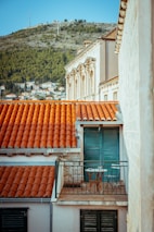 A small balcony with two chairs and a table overlooks red-tiled rooftops against the backdrop of a hilly landscape. The balcony has blue shutters and stone walls, reminiscent of Mediterranean architecture.