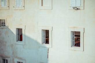 A building facade with several windows, some of which are open, revealing glimpses of interior furniture such as chairs. The wall is painted in a muted beige color, and there is a shadow cast across part of the surface.
