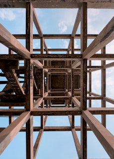 A complex structure of wooden beams forms a geometric grid pattern, viewed from below against a backdrop of a bright blue sky with scattered white clouds. The beams display a weathered texture, with an intricate interplay of light and shadow, creating a sense of depth and perspective.